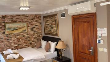 A bedroom with a brick wall, a bed with pillows, a bedside table with a lamp, a wooden door, and an air conditioning unit.