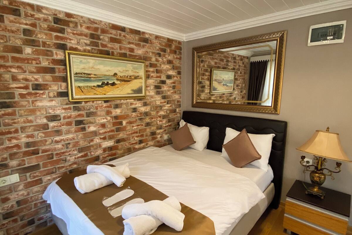A hotel room with a brick wall, a large bed with white linens, two pillows, a bedside table with a lamp, and a mirror above the bed.