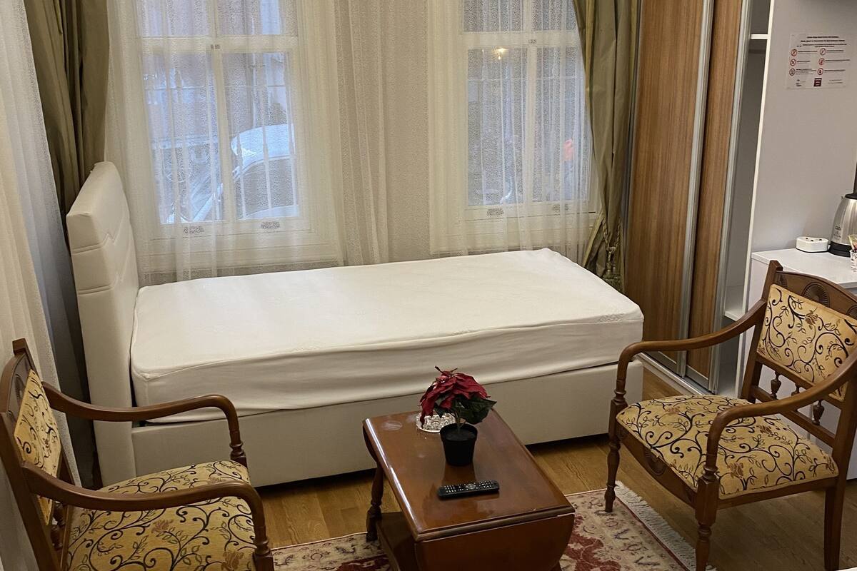 A hotel room with a bed, two chairs, a small table with a plant, a chandelier, and a television.