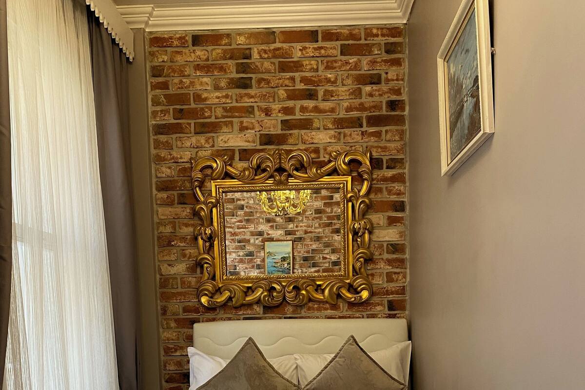 A hotel room with a brick wall, a large ornate mirror, a bed with white bedding and two pillows, and a window with sheer curtains.