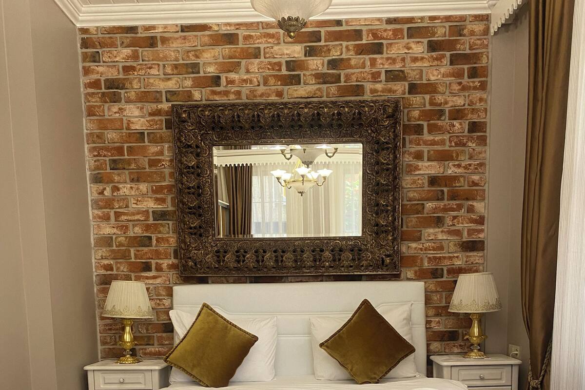 A double bed with white linens, two gold pillows, and a brown throw blanket. There are two bedside tables with lamps, a large mirror above the bed, and a brick wall behind.