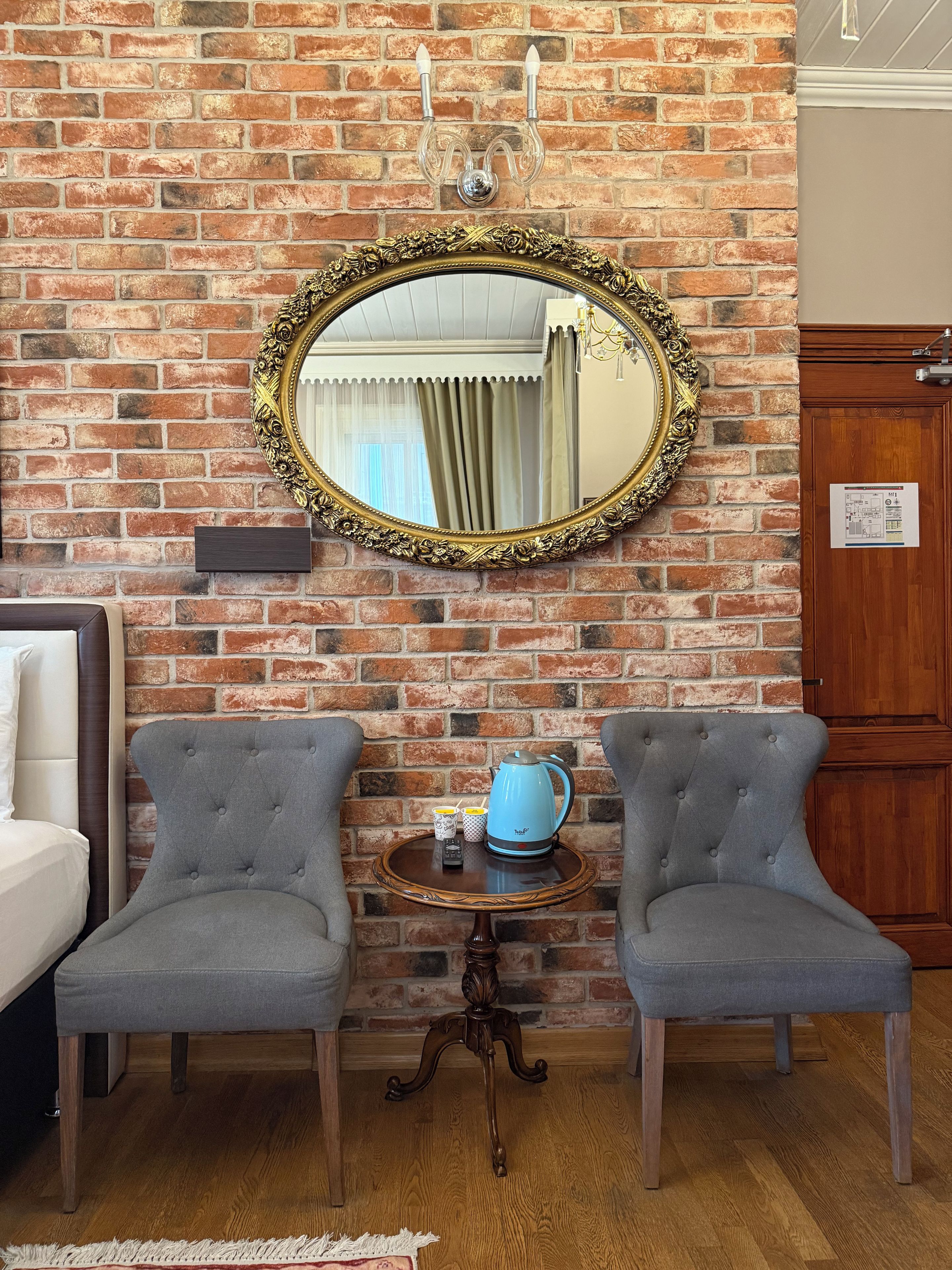 A room with a brick wall, a large ornate mirror, two grey upholstered chairs, a small round table with a blue kettle, and a wooden door.