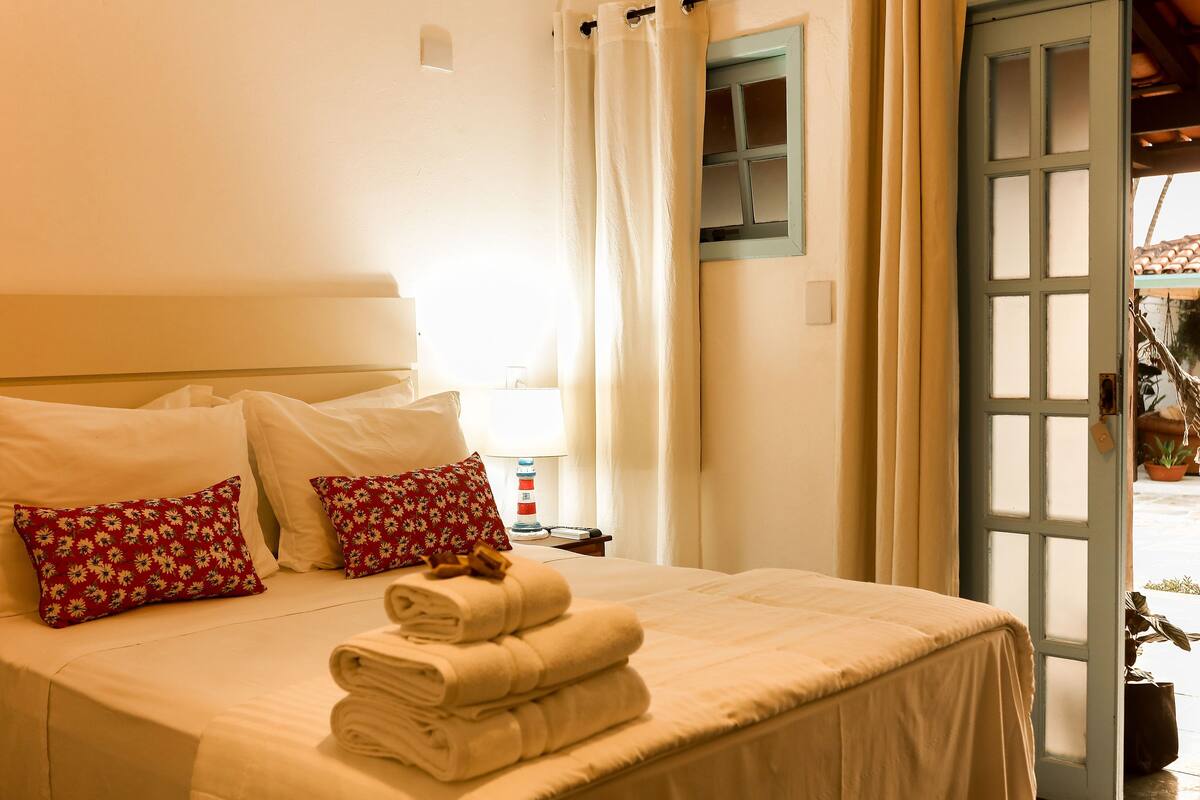 Una cama bien hecha con sábanas blancas y almohadas florales, una pila de toallas dobladas y vistas al exterior a través de una puerta abierta.