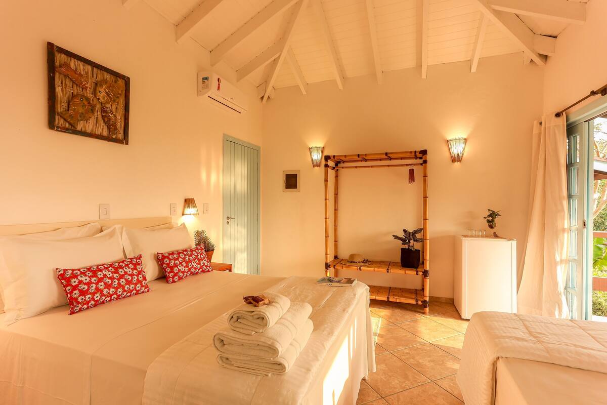 Un dormitorio con cama, almohadas, mesita de noche con lámpara, espejo y ventana con cortinas.
