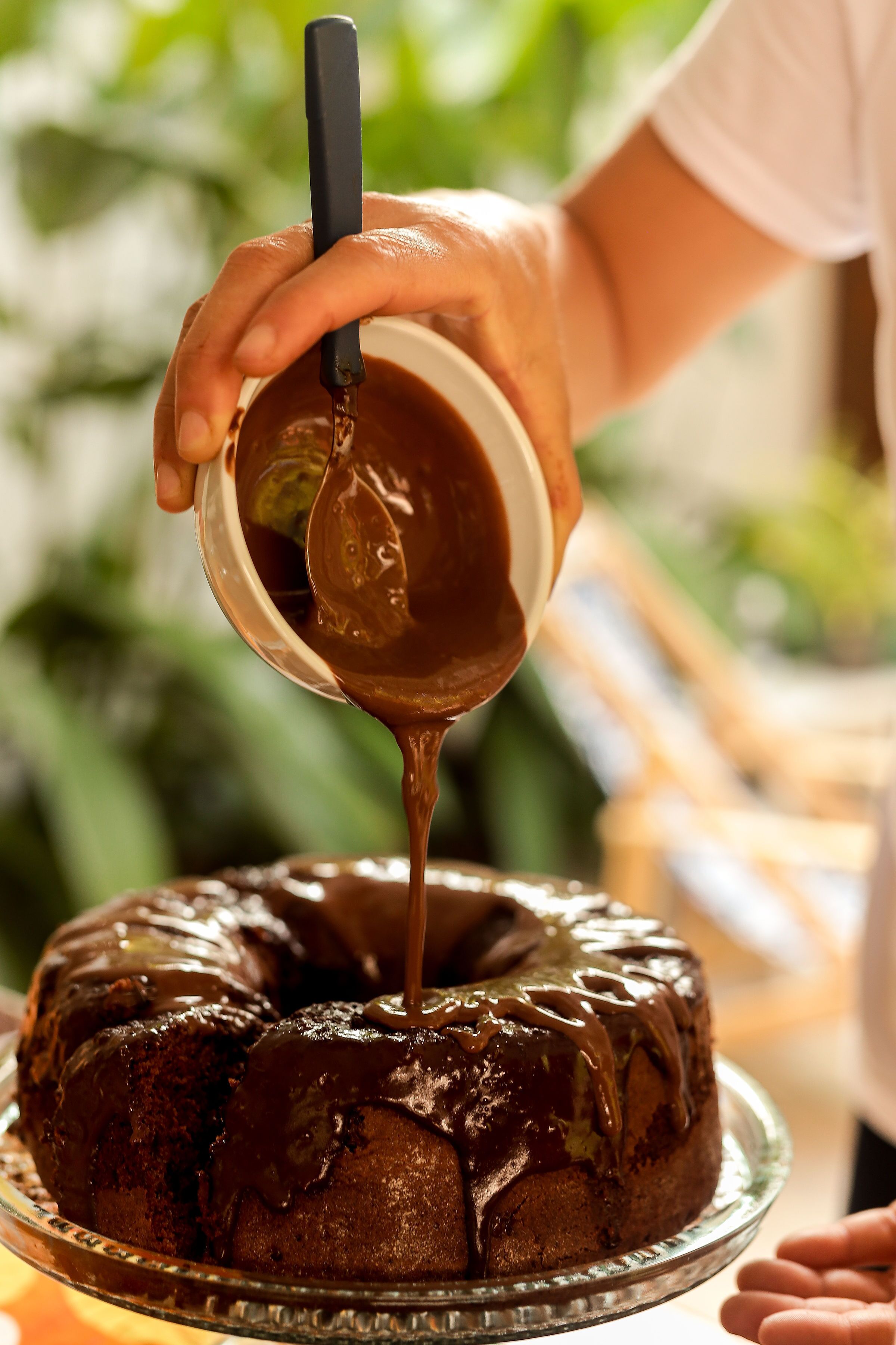 Uma pessoa despejando calda de chocolate sobre um bolo bundt em um prato de vidro.