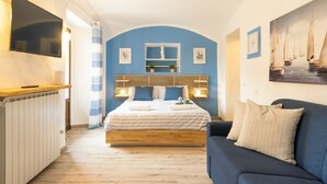 Un dormitorio con cama, pared azul, cabecera de madera, televisor, sofá y un cuadro.