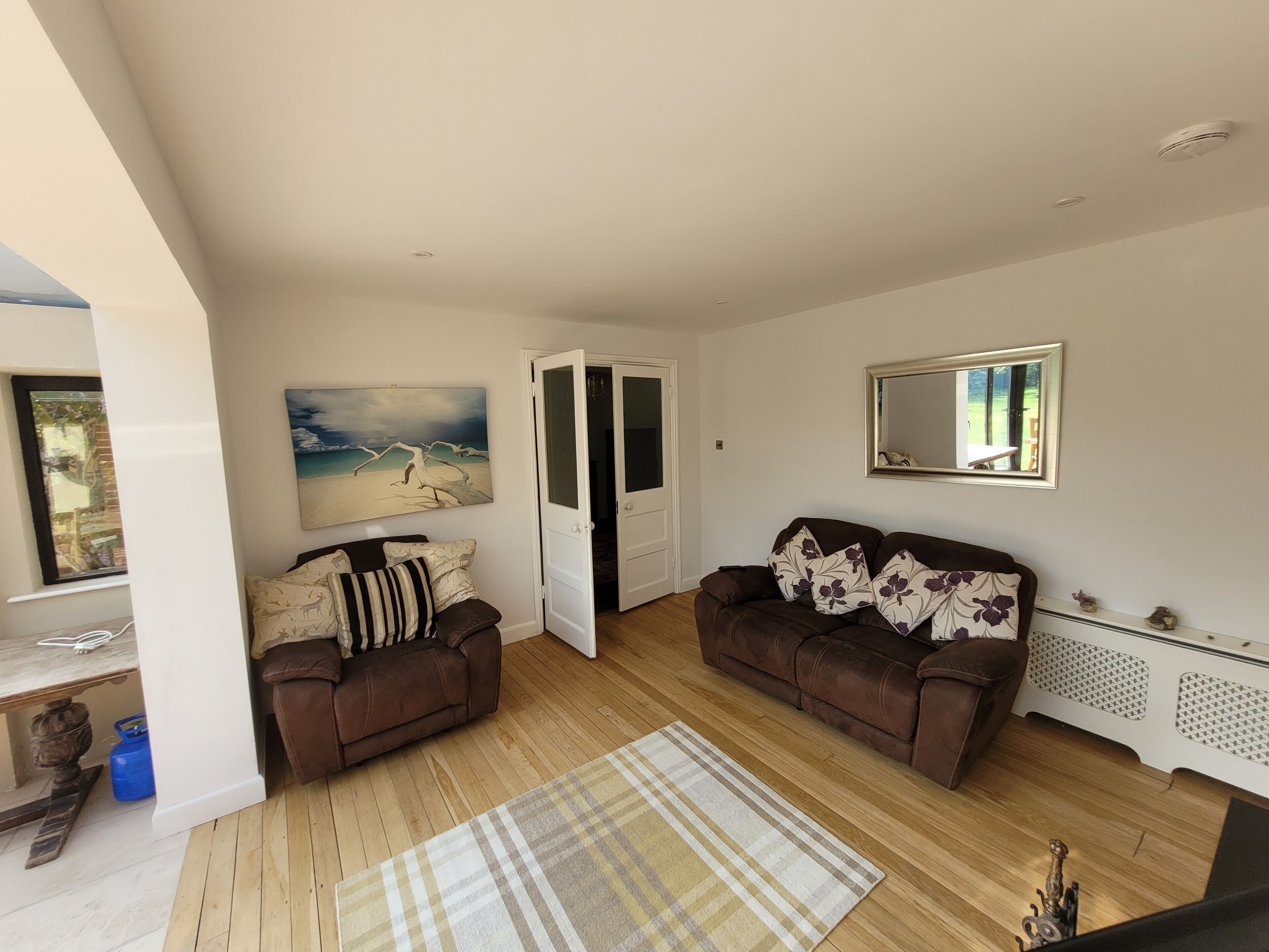 A living room with two brown sofas, a wooden floor, a painting of a beach, a mirror, and a door leading to another room.