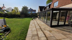 A patio with stone flooring, a glass enclosure, and a lawn. - Remarkable 8 Bed Family House Sleeping 16 w/ Pool (Farnborough)