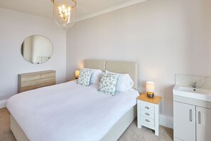 A neatly arranged bedroom with a bed, bedside tables, a mirror, and a bathroom with a sink and cabinet.