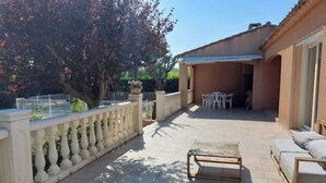 A covered patio area with a tiled floor, a wooden bench, and a table. There is a railing along the left side and a building with a tiled roof in the background. - Chez Nicolle et Jean - House near Narbonne (Canet)