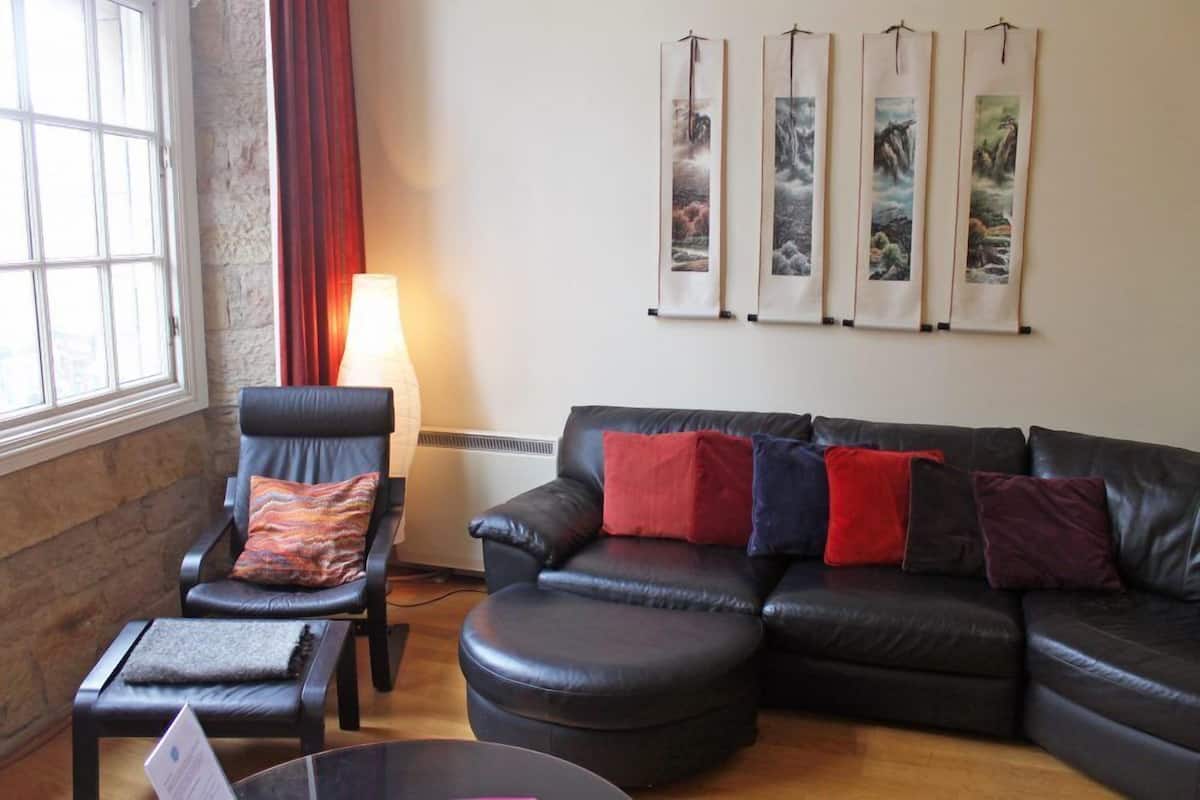 Ein Wohnzimmer mit einem schwarzen Ledersofa, einem schwarzen Sessel und einem kleinen Tisch mit einer Lampe. Vier gerahmte Kunstwerke sind an der Wand angebracht.
