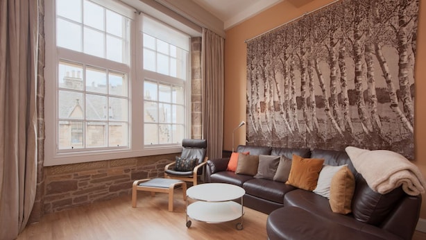 A living room with a leather sofa, a wooden coffee table, a chair, and a large window. - Royal Mile Mansions by Edinburgh City Apartments (Edinburgh)