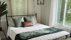 Een netjes opgemaakt bed met een bedovertrek met patroon en kussens, een hanglamp en uitzicht op groen door een raam.