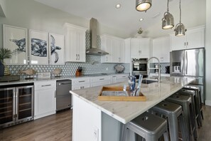 A modern kitchen with white cabinets, a marble countertop, stainless steel appliances, and a central island with stools. - Private home with sweeping water views, 2 min walk to the beach. Sleeps 13! (Navarre)