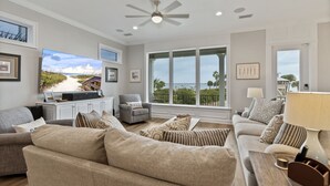 A spacious living room with a large TV, comfortable sofas, a ceiling fan, and large windows offering a view of the outdoors. - 🌊 Narrvelous – Sound-View 5BR Home w/ Pool, Tennis, Firepit & Walk to Beach (Navarre)