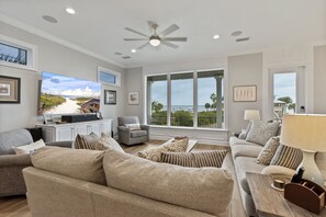 A spacious living room with a large TV, comfortable sofas, a ceiling fan, and large windows offering a view of the outdoors.
