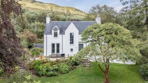 A white house with a grey roof, surrounded by lush greenery and a hill in the background. - Captivating 4-bed House in Killin (Killin)