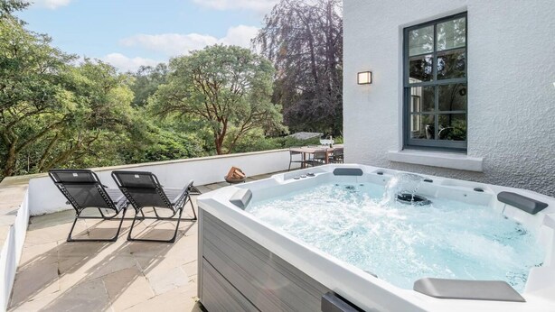 A hot tub on a patio with two lounge chairs, surrounded by trees and a white building with a window. - Captivating 4-bed House in Killin (Killin)