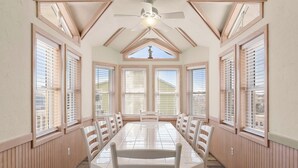 A dining room with a long table, white chairs, wooden paneled walls, and large windows with blinds. - O'Shinn View: Short Walk to Beach | Pool, Hot Tub (Corolla)