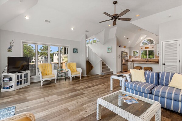 A spacious living room with a television, white wicker furniture, a striped sofa, and a staircase leading to a kitchen. - Ultra Classic: Private Pool | Hot Tub | Gym Access (Corolla)