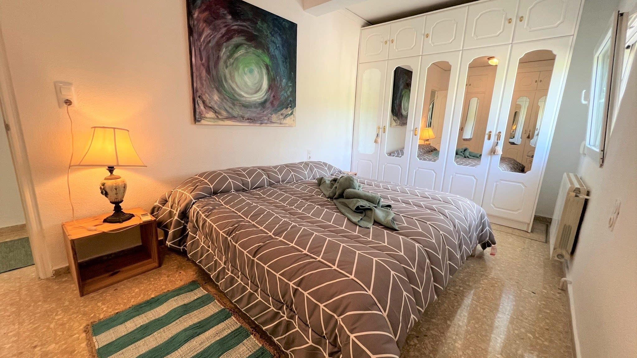 Une chambre à coucher comprenant un lit, une table de chevet, une lampe, un tableau et une armoire aux portes miroir.