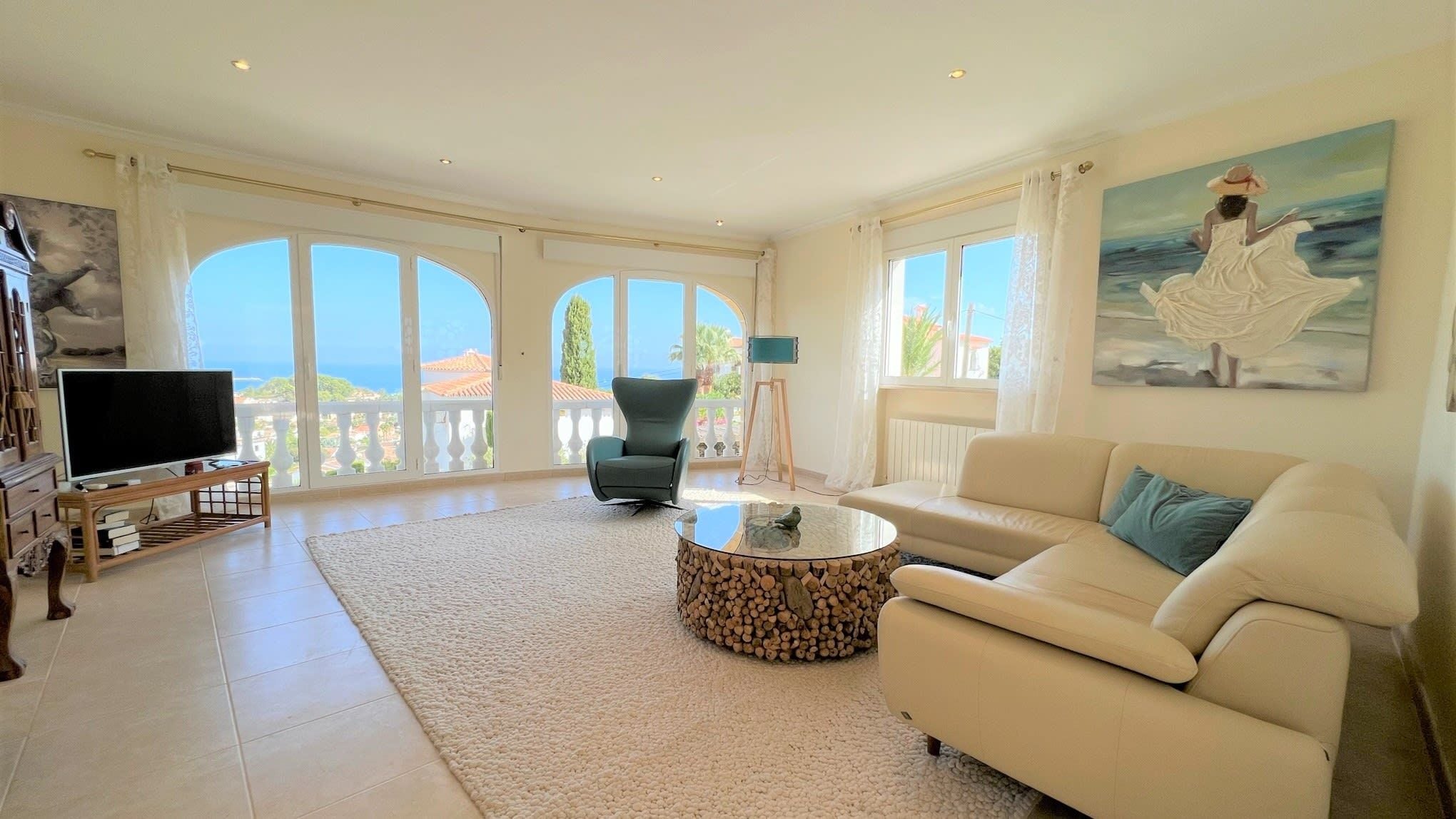 A spacious living room with a large TV, comfortable seating, and a view of the sea through large windows.