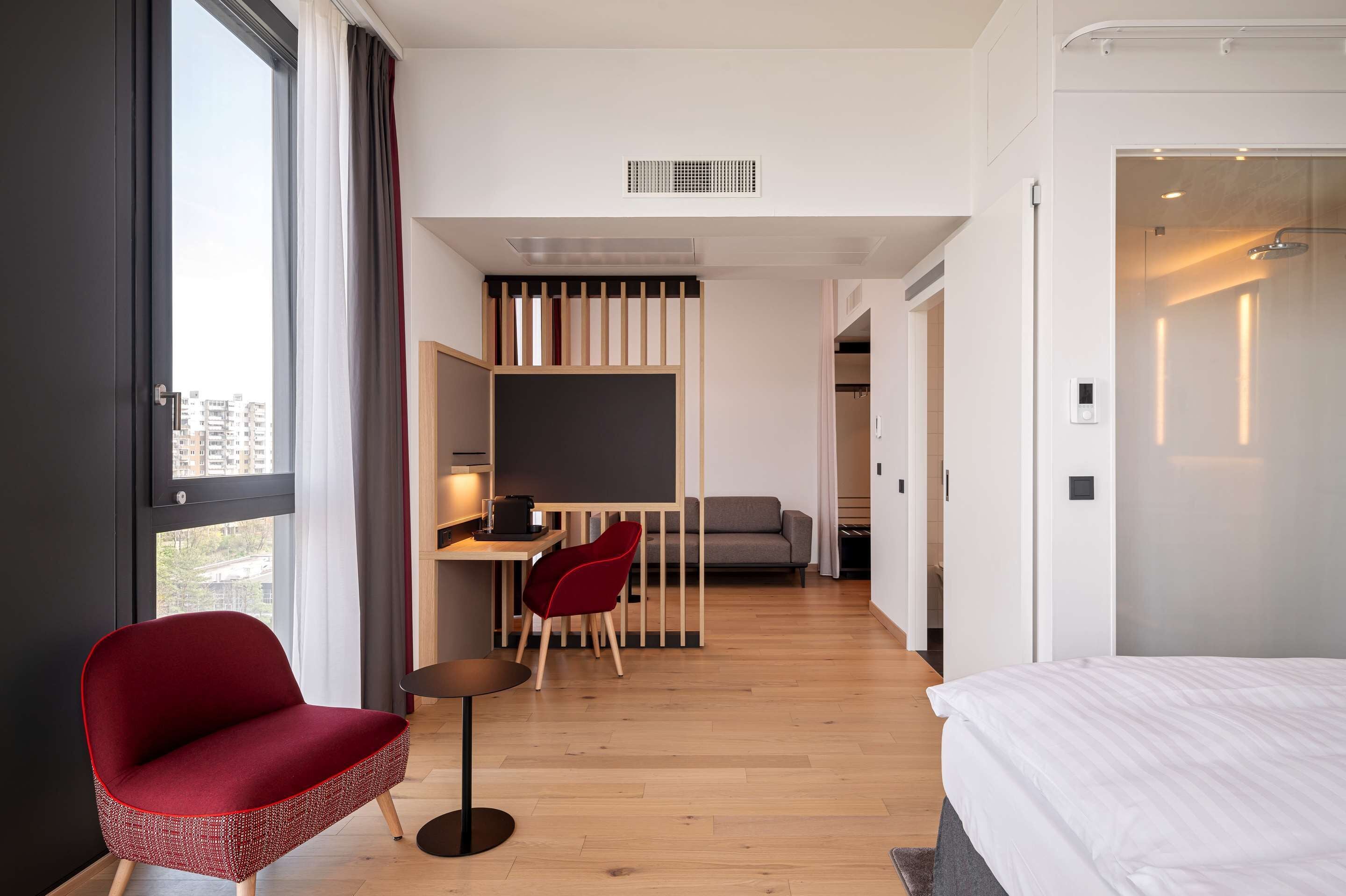 A modern hotel room with a large bed, a desk with a chair, a sofa, and a view of the city through the window.