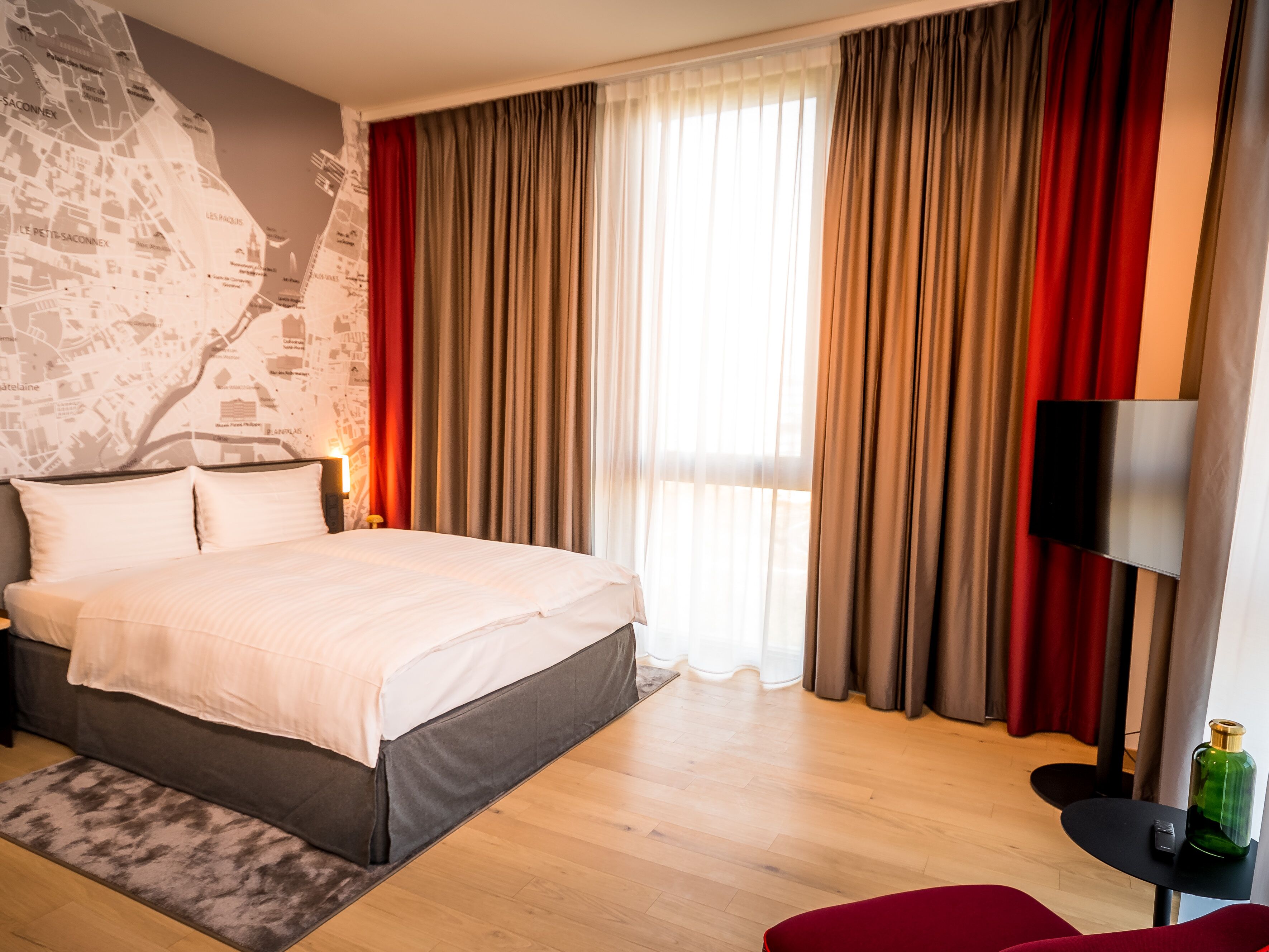 A hotel room with a bed, a map wall, a TV, and a red chair.