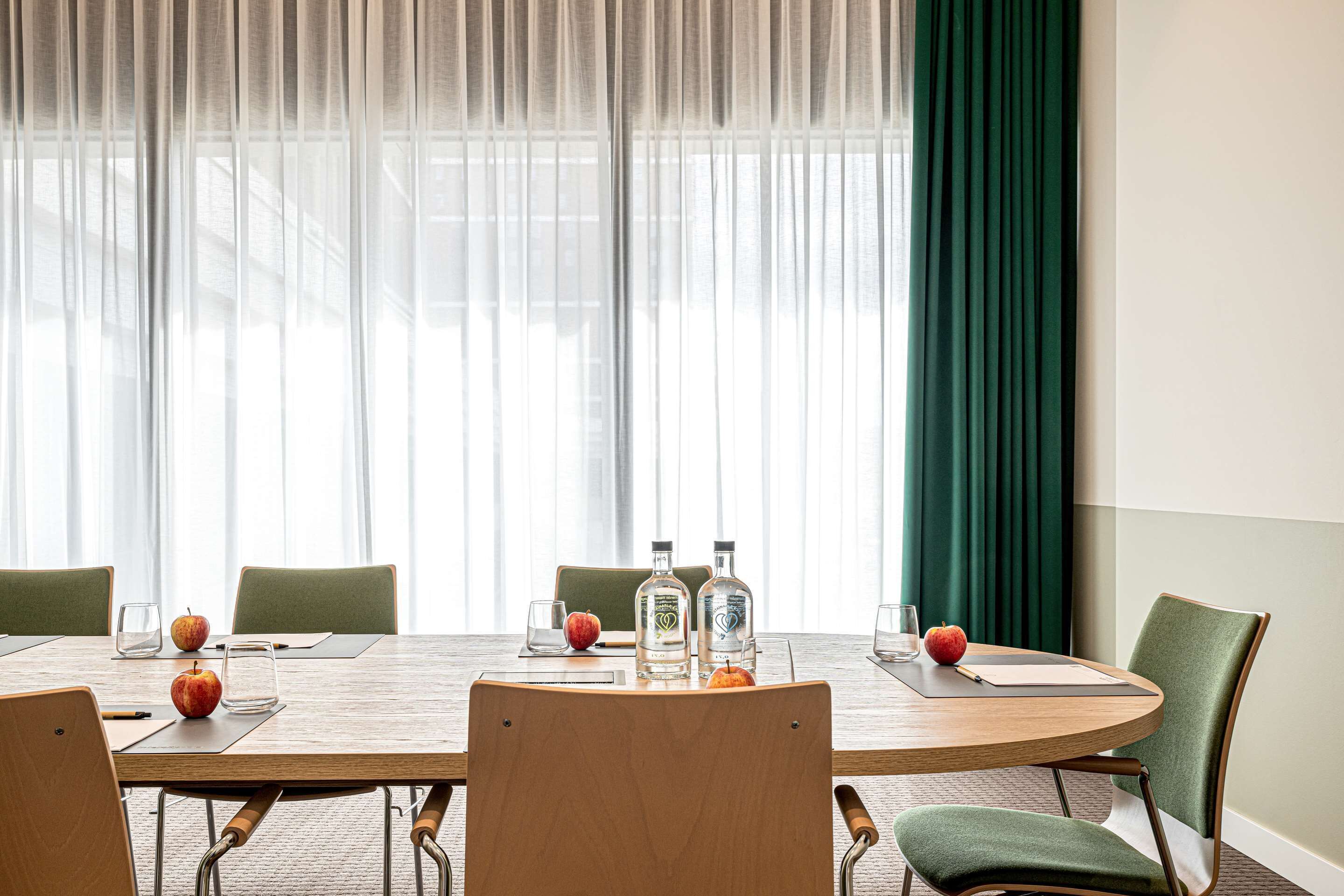 A conference room with a wooden table, green chairs, and a large window with sheer curtains.