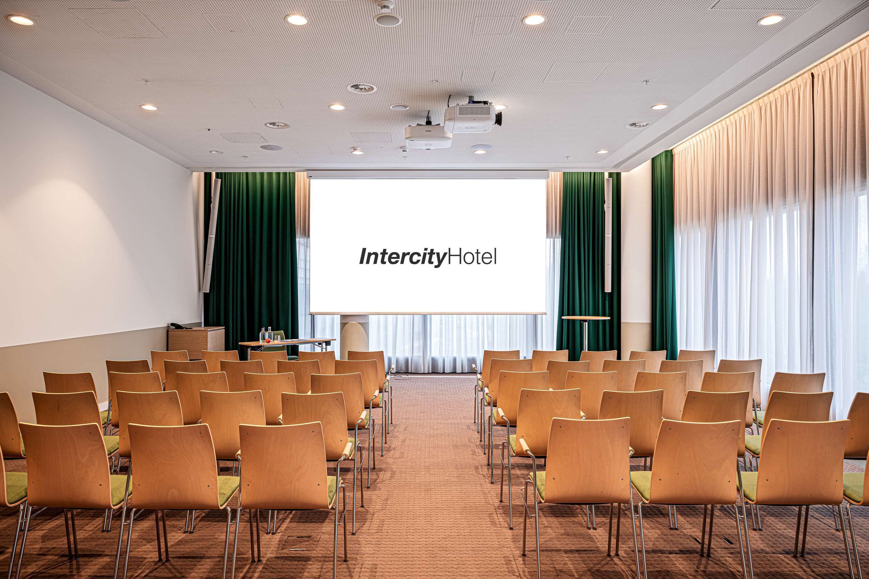 Un salón de conferencias con filas de sillas, una pantalla grande que muestra 