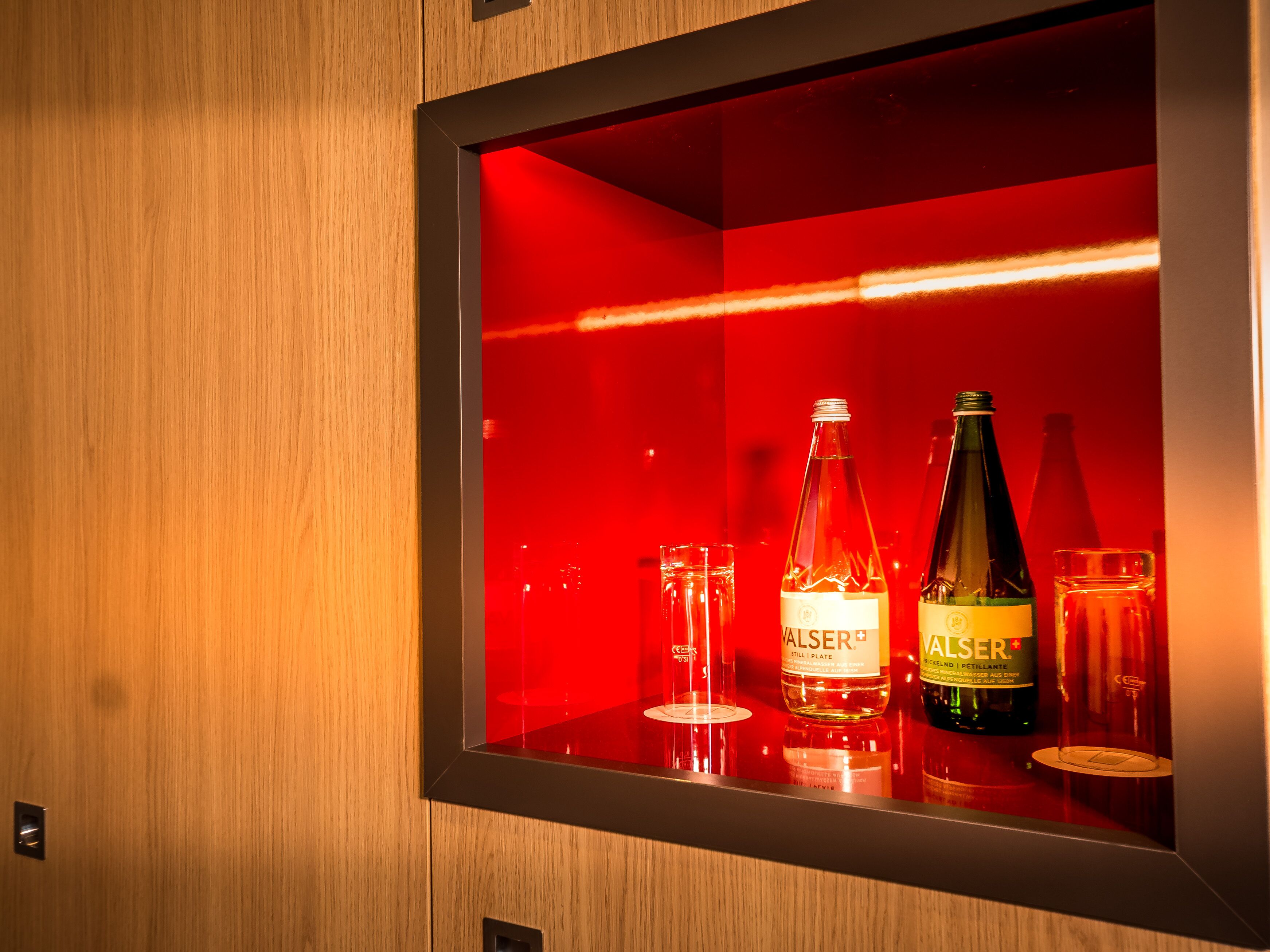 Un mini-bar à l’intérieur éclairé de lumière rouge, contenant deux bouteilles étiquetées « VALSER » et un verre.