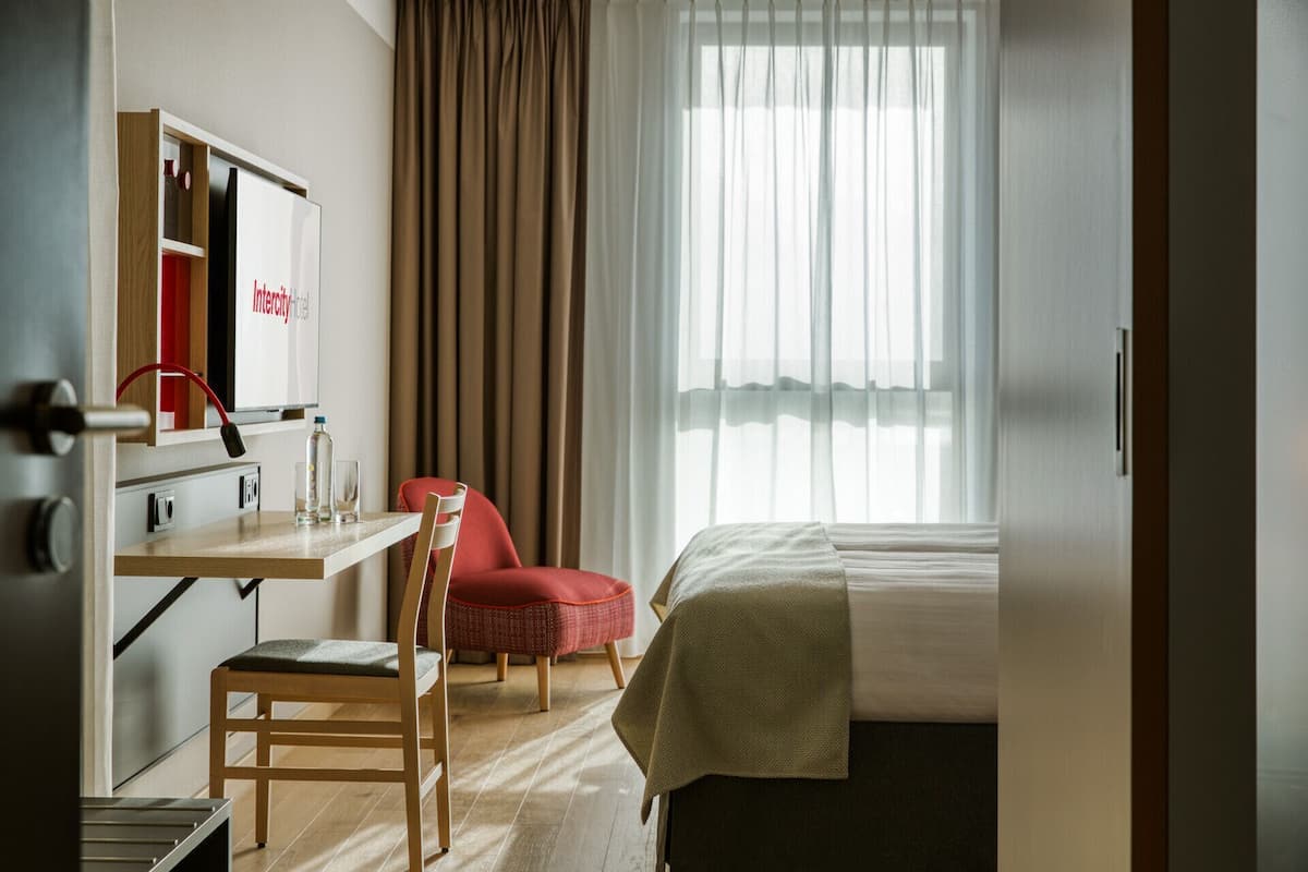 Une chambre d’hôtel avec un lit, un bureau avec une chaise, une chaise rouge, un miroir et une fenêtre avec des rideaux.