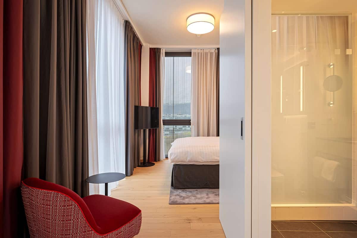 Une chambre d’hôtel avec un lit, un fauteuil rouge, une télévision et une vue sur la ville grâce à de grandes fenêtres.