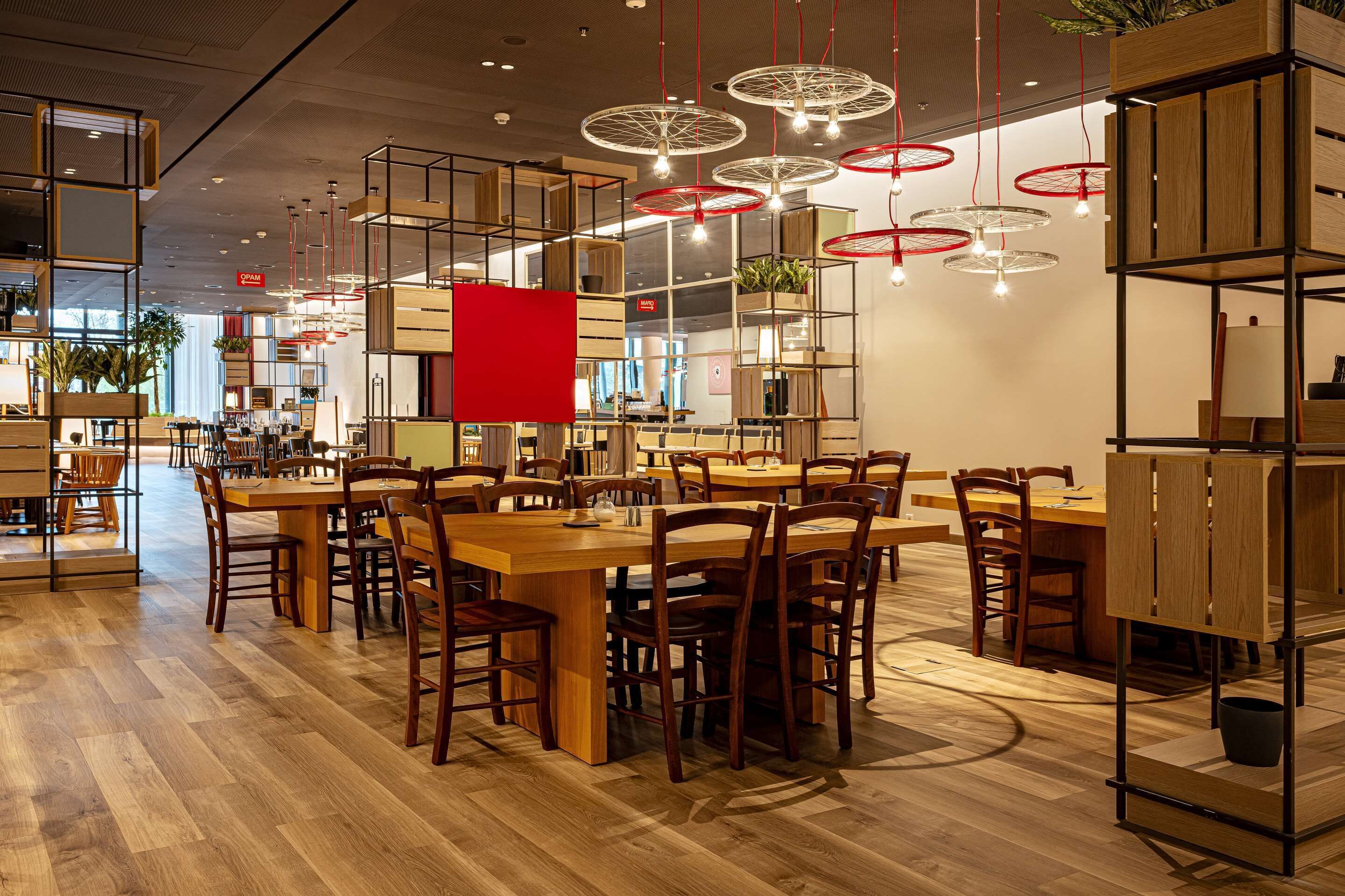 A modern dining area with wooden tables and chairs, red hanging lights, and a red accent wall.