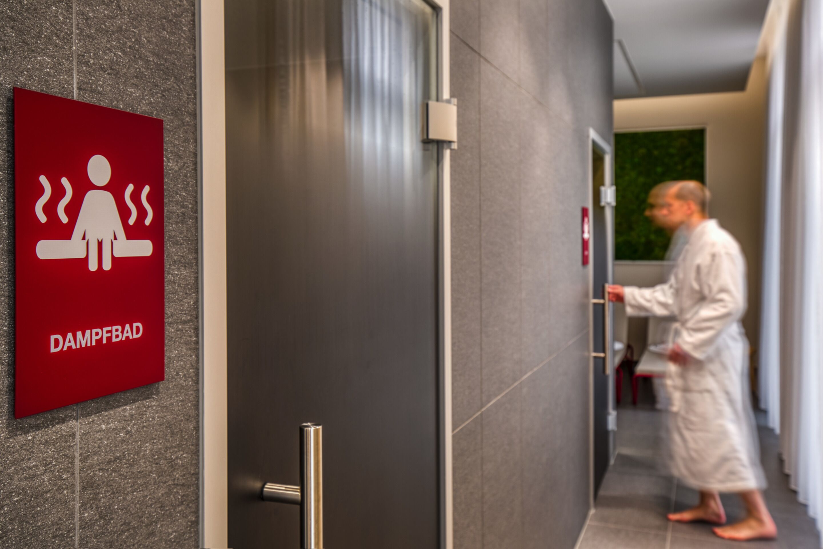 A man in a robe walking through a hallway with a red sign indicating a damp room.