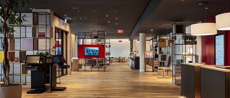 A modern hotel lobby with wooden flooring, a reception desk, a TV screen, and a seating area.