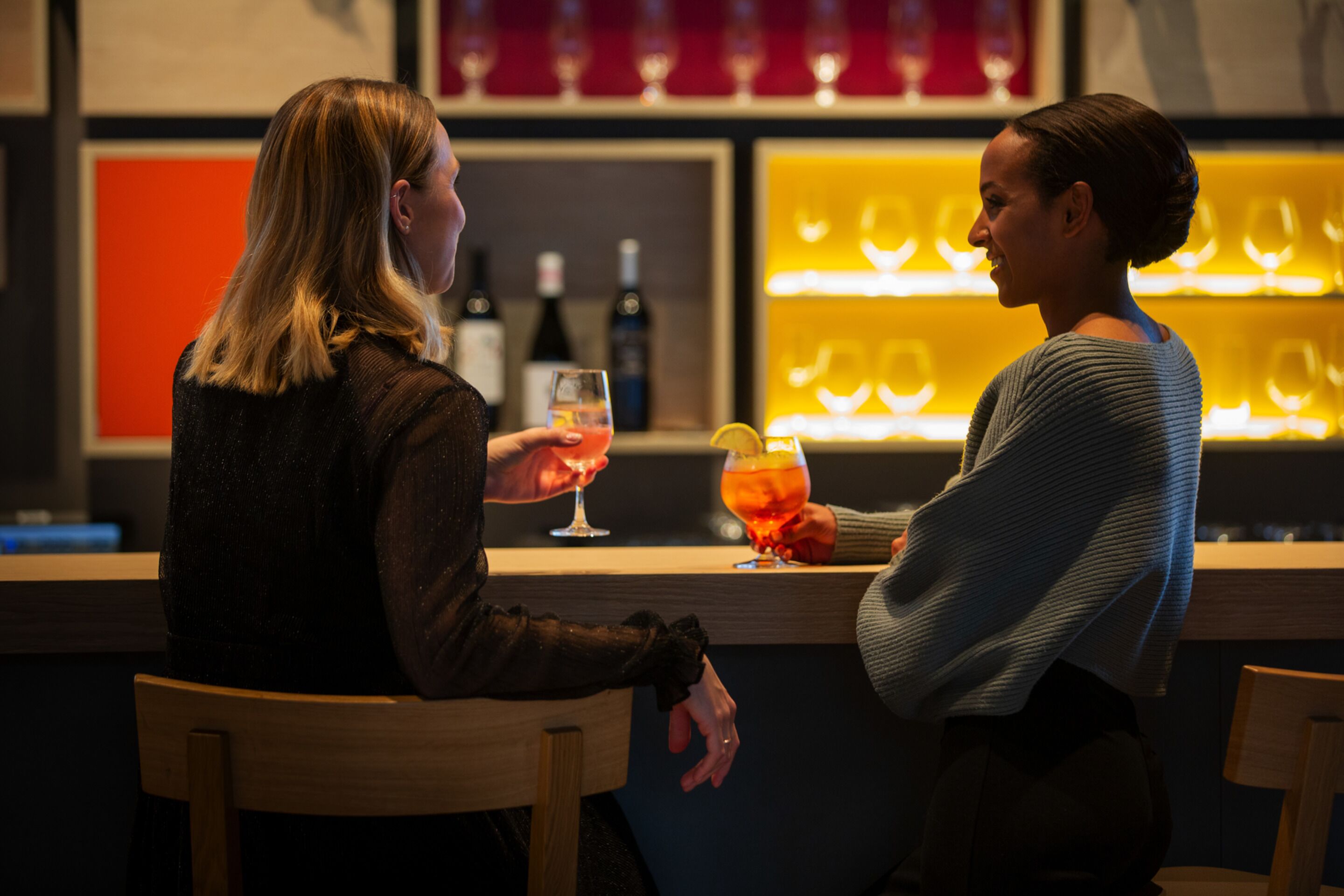 Dos personas sentadas en una barra con una bodega de vinos al fondo.
