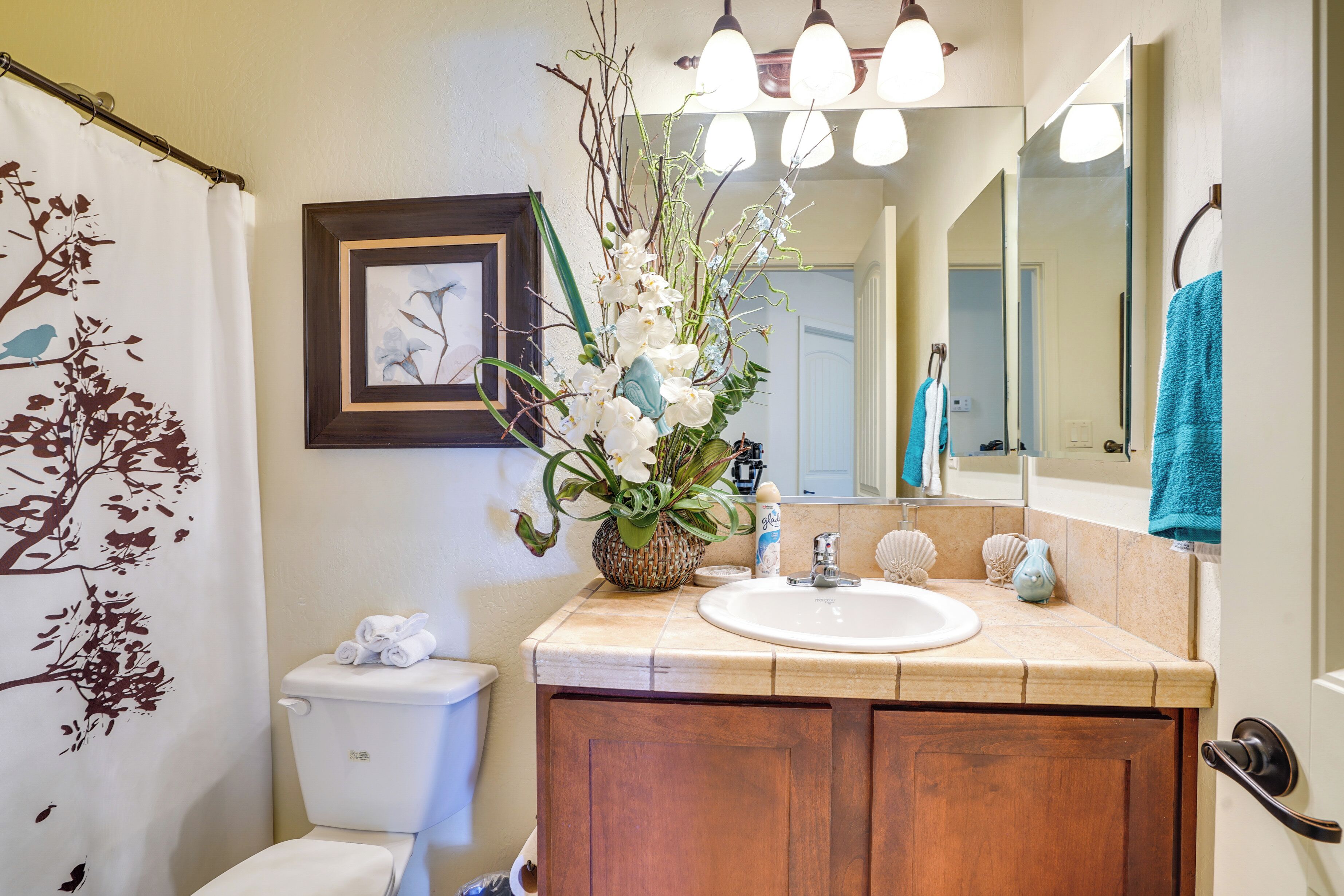 A bathroom with a toilet, sink, and mirror. There is a towel rack with a blue towel, a framed picture on the wall, and a plant on the sink counter.