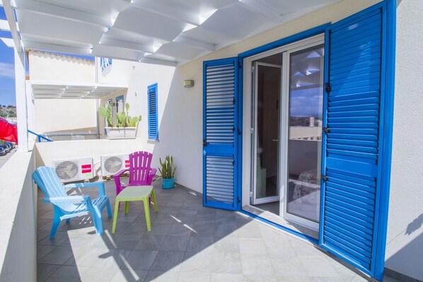 Un balcon avec des volets bleus, une table, deux chaises et un pot de fleurs.