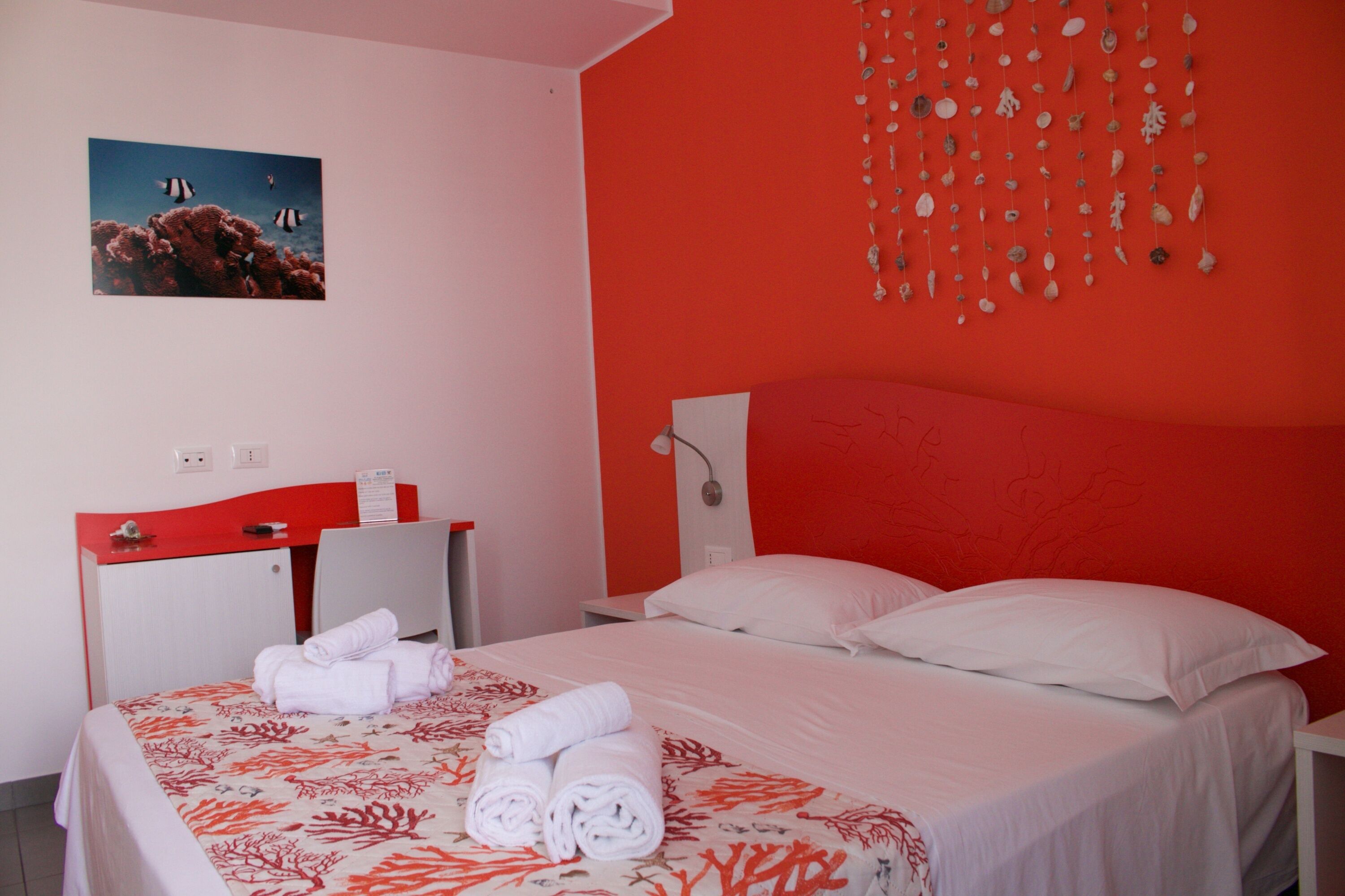 Een hotelkamer met een rode wand, een bed met witte lakens, een bureau met een rode bladplaat en een decoratie van schelpen aan de muur.