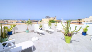 Una terraza en la azotea con sillones blancos, plantas en macetas y una vista despejada del mar y edificios a la distancia.