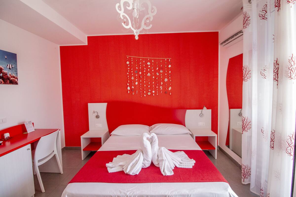 Une chambre d’hôtel avec un mur d’accent rouge, un lit avec du linge de lit blanc, un lustre, un bureau et une chaise.