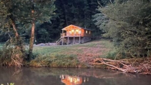 A cabin with a porch and a small deck, surrounded by trees and a body of water.