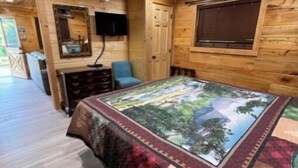 A wooden cabin interior with a bed, a ceiling fan, a TV, and a window.