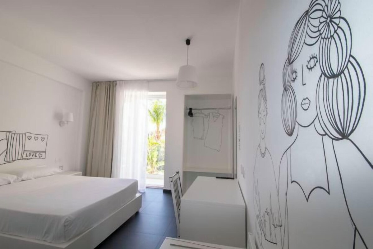 A hotel room with a bed, a desk, a chair, a hanging light fixture, and a wall with a drawing.