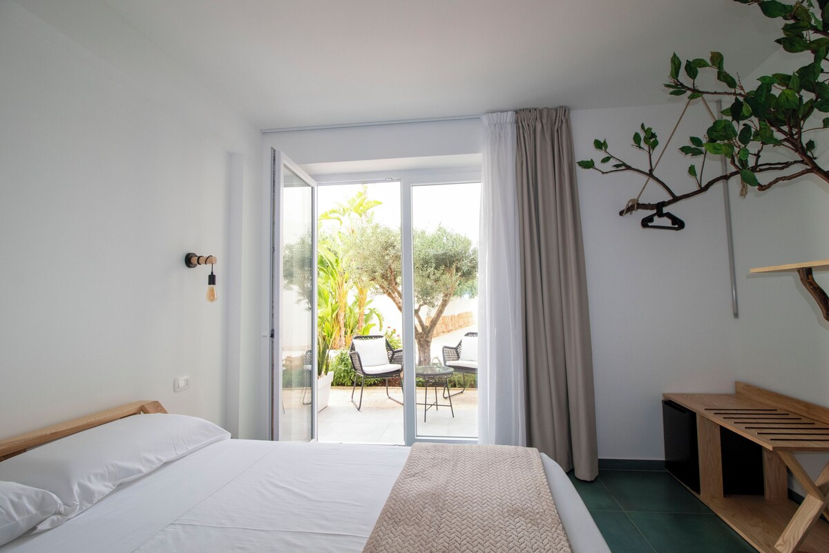 A bedroom with a bed, a wooden bench, a hanging plant, and a sliding glass door leading to an outdoor area with chairs and a table.