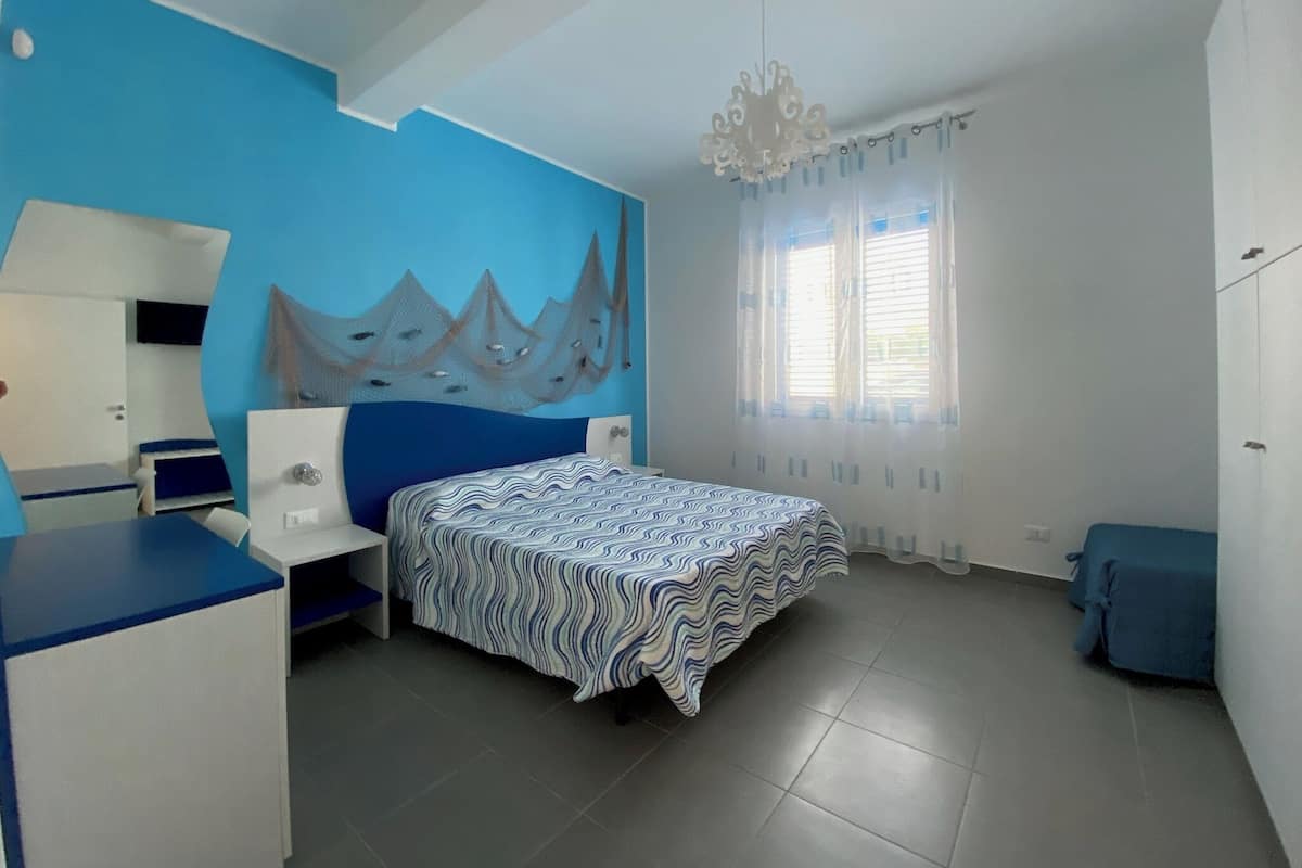 Un dormitorio con una cama, un escritorio, una silla, un espejo, una araña y una ventana con cortinas.