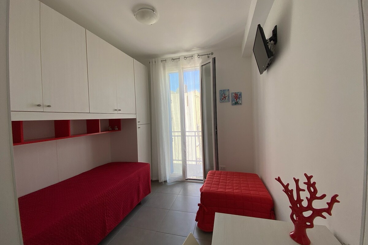 A small, well-lit room with two single beds, a TV mounted on the wall, and a window with curtains.