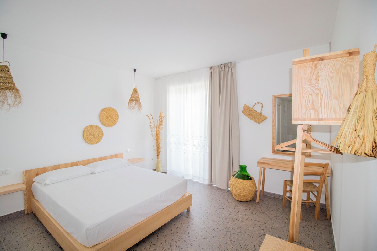 A minimalist hotel room with a wooden bed, a desk, and a large window with curtains.