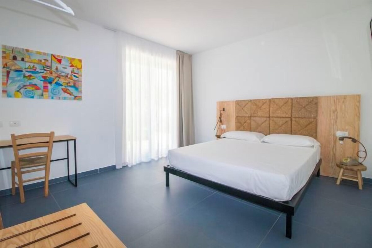 A hotel room with a bed, a desk, a chair, and a painting on the wall.